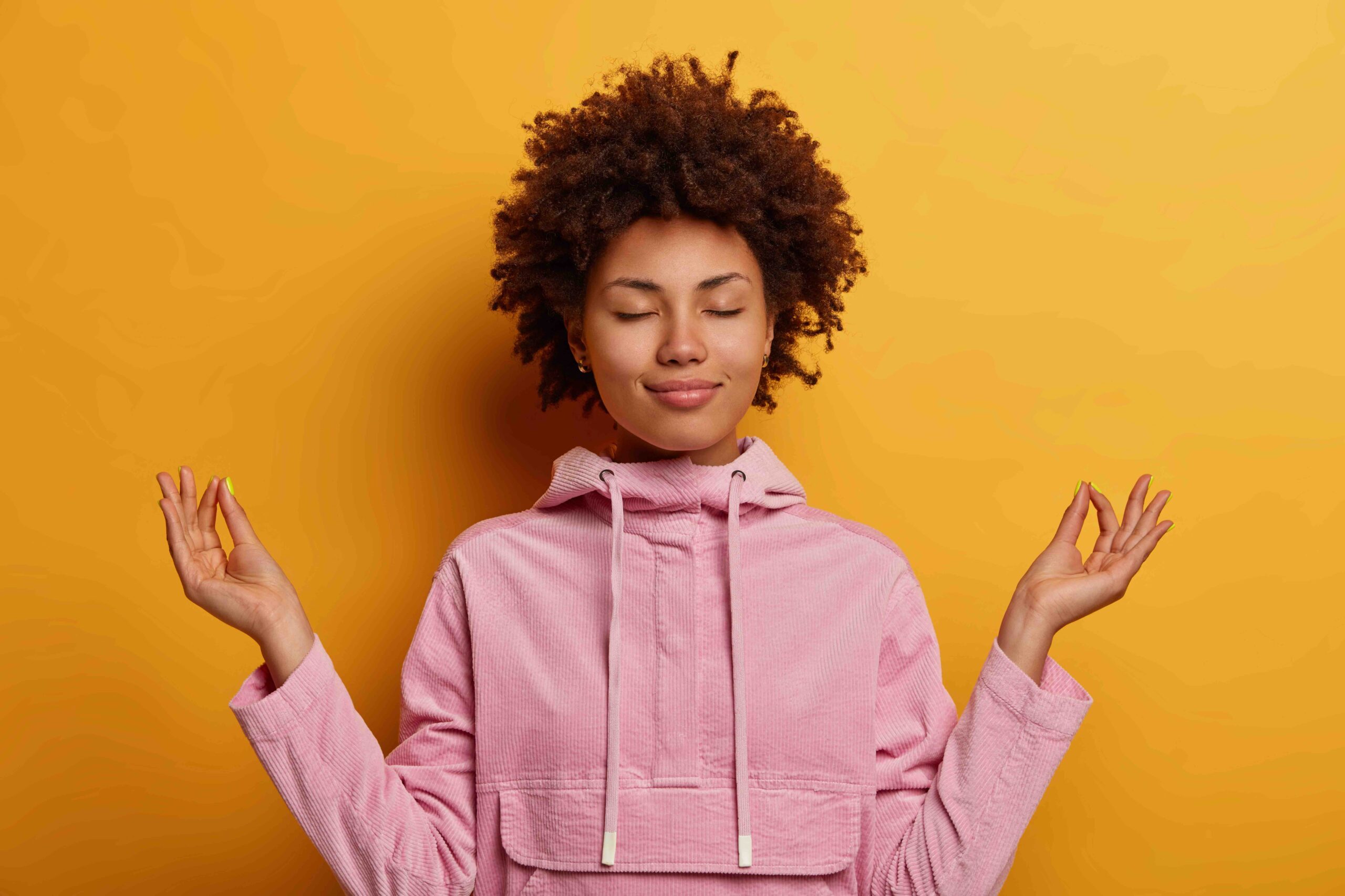 Mental Health. An ethnic woman stands in a lotus pose dressed in a pink sweatshirt on a mustard yellow background.